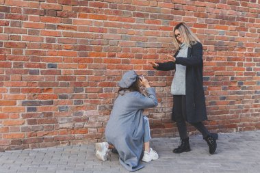 Kız, şehir sokağındaki tuğla duvarın önünde kadın arkadaşının fotoğrafını çekiyor. Fotoğrafçı ve genç şehir hayatı.