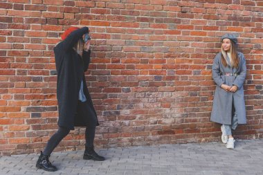 Kız, şehir sokağındaki tuğla duvarın önünde kadın arkadaşının fotoğrafını çekiyor. Fotoğrafçı ve genç şehir hayatı.
