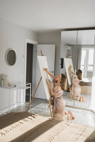 Young woman artist painting on canvas on the easel at home in bedroom - art and creativity