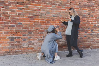 Kız, kız arkadaşının fotoğrafını tuğla duvarın önünde çekiyor.