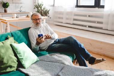 Son sınıf hipster 'ı cep telefonuyla mobil uygulama üzerinden müzik dinlerken kullanıyor. Yaşlı erkek video akıllı telefon izlerken yüz koruyucu maske takıyor.