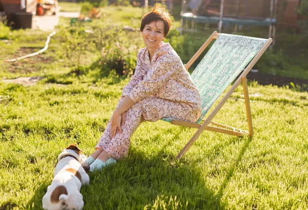 Young woman in pajama is resting in chair on a green lawn on sunny summer day - village and country life concept