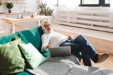 Kulaklıklı yaşlı bir adamın portresi. Evde dizüstü bilgisayardan müzik dinliyor. Aktif yaşlılar.