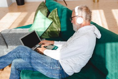 Kapüşonlu ve gözlüklü yakışıklı adam evde kanepede oturmuş müzik dinlerken dizüstü bilgisayar kullanıyor ve gülümsüyor.