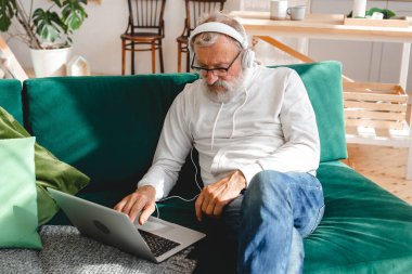 Kapüşonlu ve gözlüklü yakışıklı adam evde kanepede oturmuş müzik dinlerken dizüstü bilgisayar kullanıyor ve gülümsüyor.