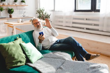 Yaşlı adam, havalı sakallı yaşlı adam video görüşmesi için cep telefonu kullanıyor. Mutluluk, yaşlılar yaşam tarzı kavramı. Akıllı telefondan çocuklara el sallayıp sohbet eden bir video. Modern.