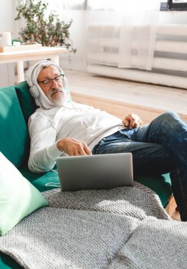 Kulaklıklı yaşlı bir adamın portresi. Evde dizüstü bilgisayardan müzik dinliyor. Aktif yaşlılar.