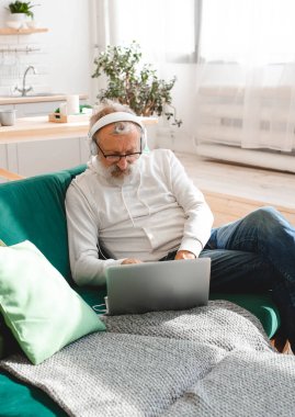 Kapüşonlu ve gözlüklü yakışıklı adam evde kanepede oturmuş müzik dinlerken dizüstü bilgisayar kullanıyor ve gülümsüyor.