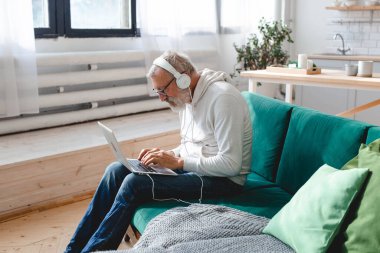Kapüşonlu ve gözlüklü yakışıklı adam evde kanepede oturmuş müzik dinlerken dizüstü bilgisayar kullanıyor ve gülümsüyor.