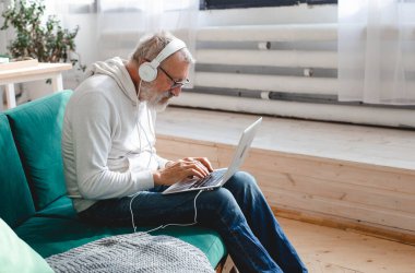 Kapüşonlu ve gözlüklü yakışıklı adam evde kanepede oturmuş müzik dinlerken dizüstü bilgisayar kullanıyor ve gülümsüyor.