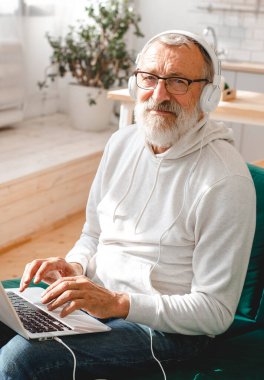 Kapüşonlu ve gözlüklü yakışıklı adam evde kanepede oturmuş müzik dinlerken dizüstü bilgisayar kullanıyor ve gülümsüyor.