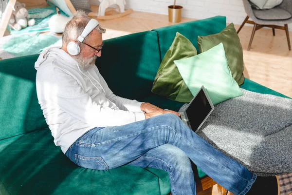 Kapüşonlu ve gözlüklü yakışıklı adam evde kanepede oturmuş müzik dinlerken dizüstü bilgisayar kullanıyor ve gülümsüyor.