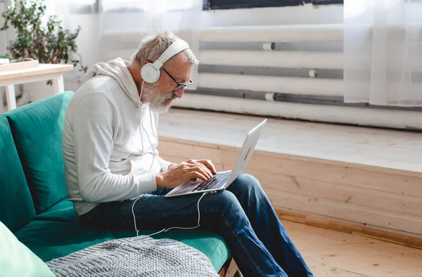 Kapüşonlu ve gözlüklü yakışıklı adam evde kanepede oturmuş müzik dinlerken dizüstü bilgisayar kullanıyor ve gülümsüyor.