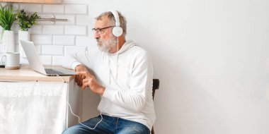 Kapüşonlu ve gözlüklü yakışıklı adam evde kanepede oturmuş müzik dinlerken dizüstü bilgisayar kullanıyor ve gülümsüyor.