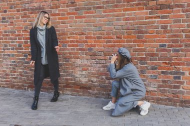Kız, şehir sokağındaki tuğla duvarın önünde kadın arkadaşının fotoğrafını çekiyor. Fotoğrafçı ve genç şehir hayatı.