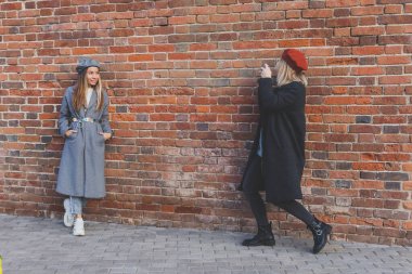 Kız, şehir sokağındaki tuğla duvarın önünde kadın arkadaşının fotoğrafını çekiyor. Fotoğrafçı ve genç şehir hayatı.
