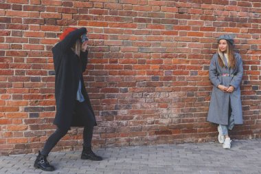 Kız, kız arkadaşının fotoğrafını tuğla duvarın önünde çekiyor.