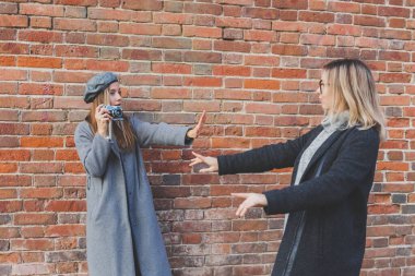 Kız, kız arkadaşının fotoğrafını tuğla duvarın önünde çekiyor.