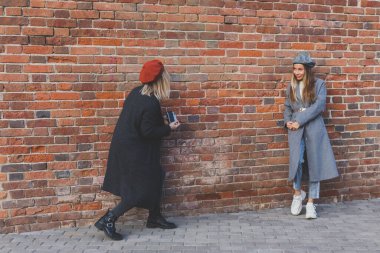 Kız, kız arkadaşının fotoğrafını tuğla duvarın önünde çekiyor.