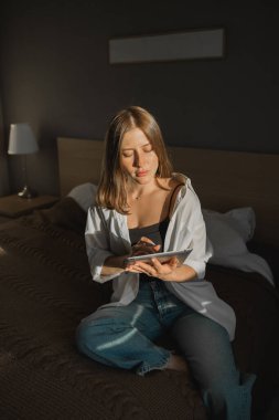 Pencerenin yanında dijital tabletli gölgesi olan güzel bir kadının portresi. Sabah ilkbahar estetiği ve sosyal ağlar dijital teknolojiler