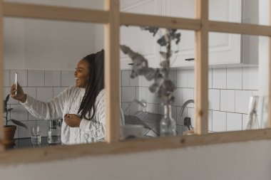 Happy modern African American young girl talking on video chat and sit home communication concept. Smiling hipster girl making self portrait on her telephone at home