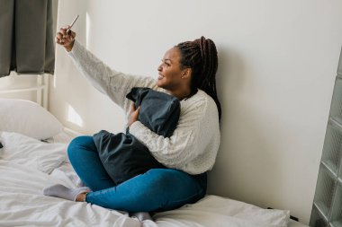 Happy modern African American young girl talking on video chat and sit home communication concept. Smiling hipster girl making self portrait on her telephone at home