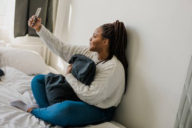 Happy modern African American young girl talking on video chat and sit home communication concept. Smiling hipster girl making self portrait on her telephone at home