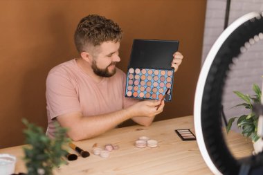 Makyaj malzemelerini elinde tutarak videosunu kaydeden bir adam. Adam blogu için kozmetik ürünü videosu yapıyor..