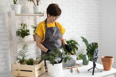 Female blogger sits in front of smartphone camera on tripod records instructional tutorial video for her blog shoots process of replanting flowers and green plants