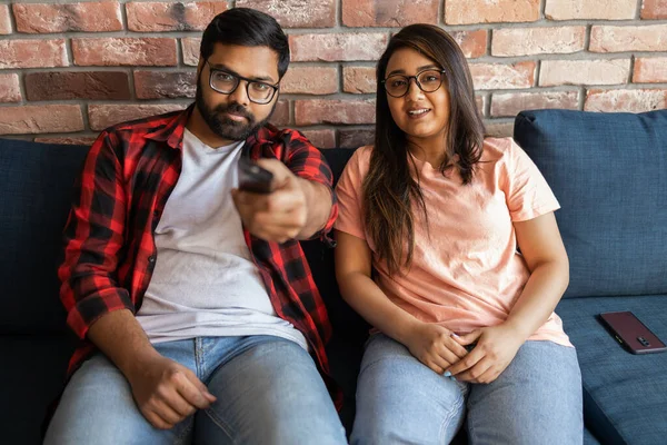 Happy indian or latino couple sitting on sofa and watching action movie on TV or criminal blockbuster on streaming service and talking and discussing acting. Girlfriend and boyfriend resting at home
