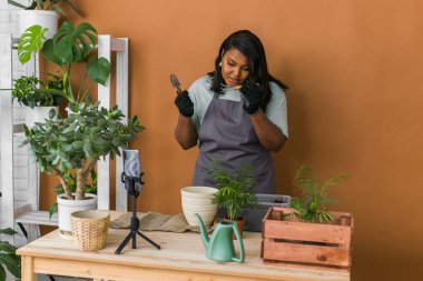 African american girl vlogger influencer or SME owner work on home video camera selfie shoot filming take care home plants and transplanting plant in flowerpot. Home gardening and florist