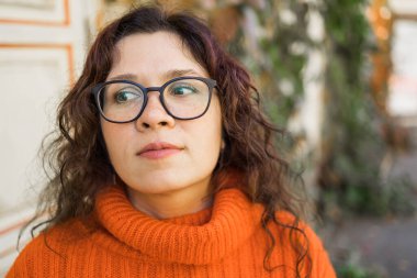 Autumn portrait of an attractive young woman in stylish glasses in a knitted fashionable orange sweater on background leaves. Girl walks in city