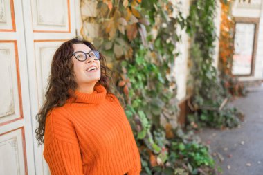 Autumn portrait of an attractive young woman in stylish glasses in a knitted fashionable orange sweater on background leaves. Girl walks in city