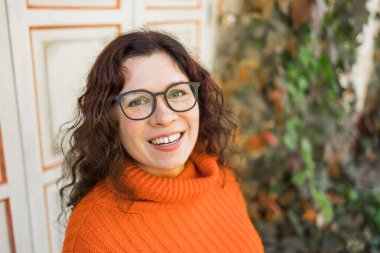 Autumn portrait of an attractive young woman in stylish glasses in a knitted fashionable orange sweater on background leaves. Girl walks in city