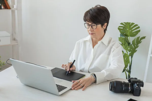 Middle aged woman graphic designer or photographer using digital graphic tablet while working at modern office, professional female retoucher sitting at modern workspace, generic design tablet and