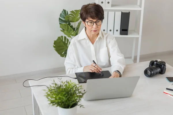 Middle aged woman graphic designer or photographer using digital graphic tablet while working at modern office, professional female retoucher sitting at modern workspace, generic design tablet and