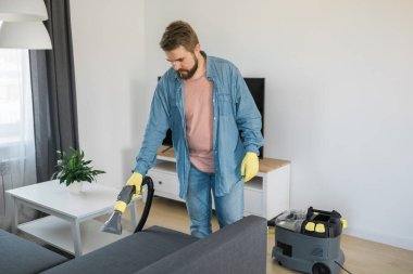 Man in protective rubber glove cleaning sofa with professionally extraction method with washing vacuum cleaner. Early spring regular cleanup. Commercial cleaning company