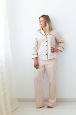 Gülümseyen sarışın kadın pijamayla oturma odasında rahat bir koç üzerinde oturuyor. Ev kıyafetleri ve evde boş zaman tatili.