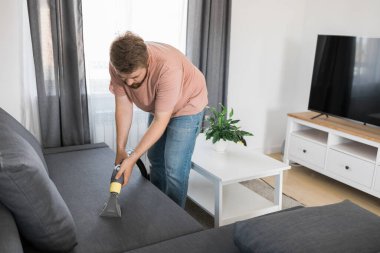 Man holding modern washing vacuum cleaner and cleaning dirty sofa with professionally detergent. Professional springclean at home