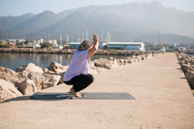 Sabahları deniz kenarında kıdemli bir kadın esneme pozisyonu alıyor. Yaşlı kadın plajda yoga yapıyor..