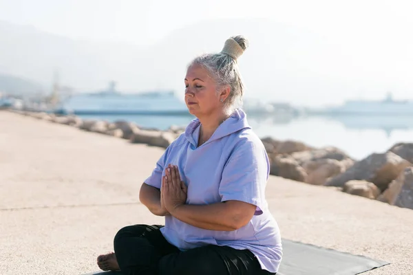 stock image Senior woman in stretching position by the sea at morning. Elderly woman doing yoga on beach.