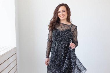 Çekici, esmer, uzun saçlı, şık, siyah elbiseli, gülümseyen ve poz veren bir kadın. Arka planda beyaz duvara karşı duran gülen kadın model.