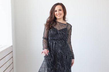 Beyaz duvarda poz veren şık, siyah parti elbiseli, baştan çıkarıcı bir kadın. Moda portresi. Doğum gününü kutluyoruz..