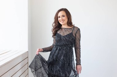 Beyaz duvarda poz veren şık, siyah parti elbiseli, baştan çıkarıcı bir kadın. Moda portresi. Doğum gününü kutluyoruz..