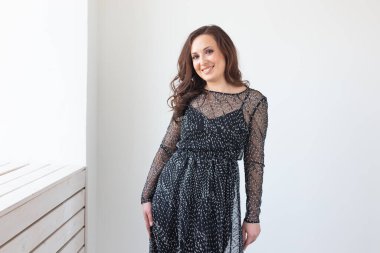 Çekici, esmer, uzun saçlı, şık, siyah elbiseli, gülümseyen ve poz veren bir kadın. Arka planda beyaz duvara karşı duran gülen kadın model.