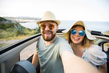 Mutlu ve güzel bir çift tatilde üstü açık bir arabayla selfie çektiriyorlar. Kiralık arabalar ve tatil konsepti