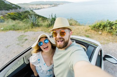 Mutlu ve güzel bir çift tatilde üstü açık bir arabayla selfie çektiriyorlar. Kiralık arabalar ve tatil konsepti