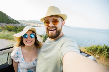 Mutlu ve güzel bir çift tatilde üstü açık bir arabayla selfie çektiriyorlar. Kiralık arabalar ve tatil konsepti