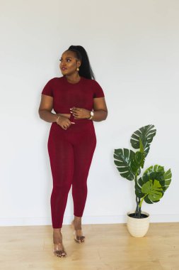 Beyaz duvarın önünde duran şişman, Afro-Amerikan kadının portresi. Kırmızı kısa tişört ve tayt giyen bir kadın. Spor giysileri ve ev giysileri.