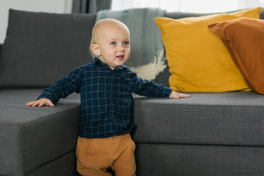 Küçük çocuk gülüyor. Oturma odasındaki kanepenin yanında durup eğleniyor. Sevimli bebek ilk adımlarını yalnız atıyor. Mutlu çocukluk ve çocuk bakımı